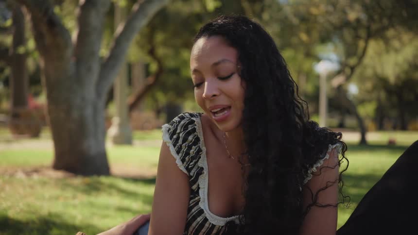 A beautiful young woman with long curly hair smiles shyly while talking with her partner in a sunlit park