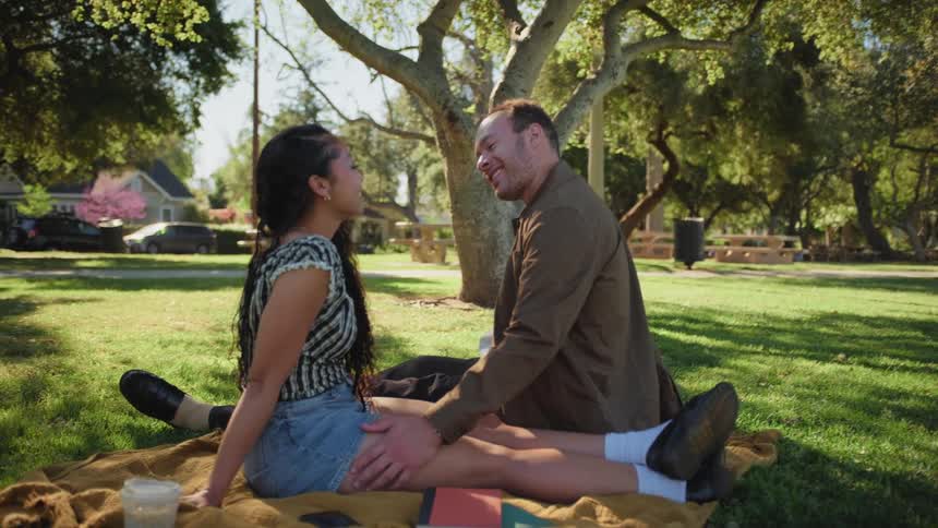 An affectionate interracial couple sits on a blanket in a sunny park, smiling and caressing each other during a romantic picnic date
