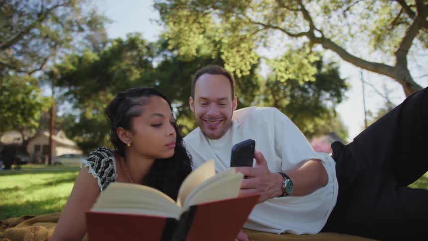 A happy couple enjoys a relaxing afternoon in the park, reading a book and sharing a funny moment on a smartphone