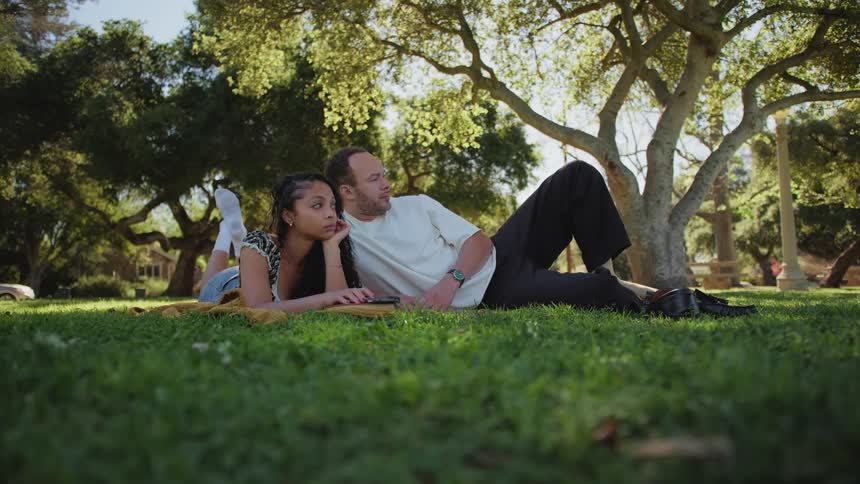 A young man and woman lie together on a blanket on the green grass, relaxing in a sunlit park