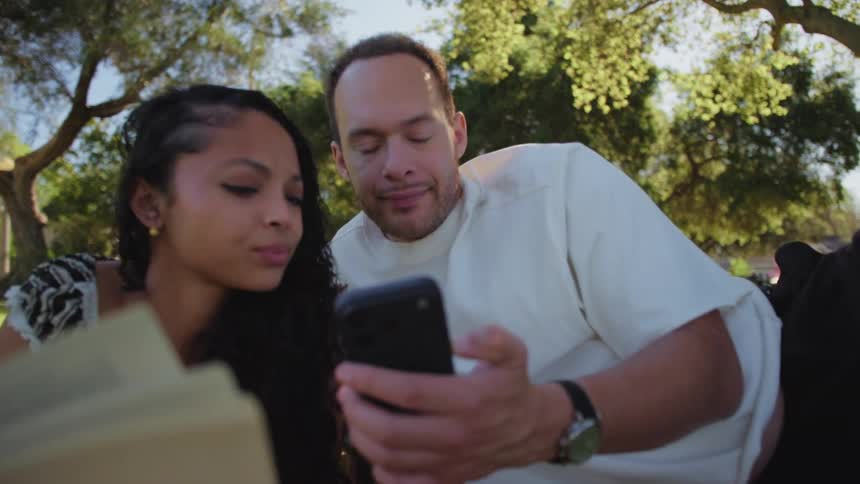 A young couple lying on a blanket in a park look at a phone together with serious expressions