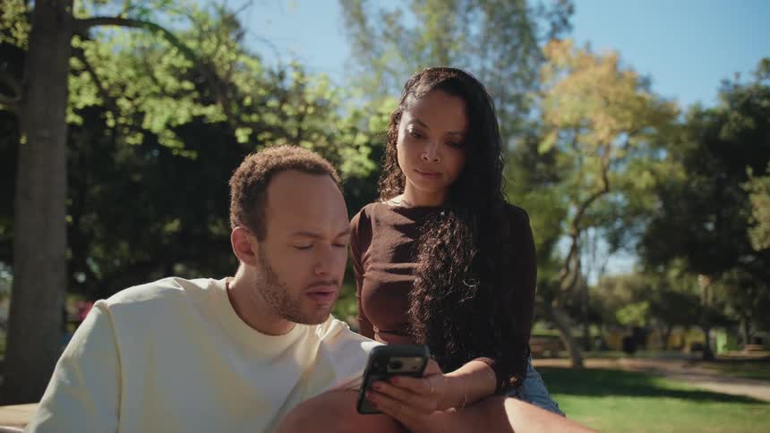 A young couple sits close together in a sunny park looking at a smartphone