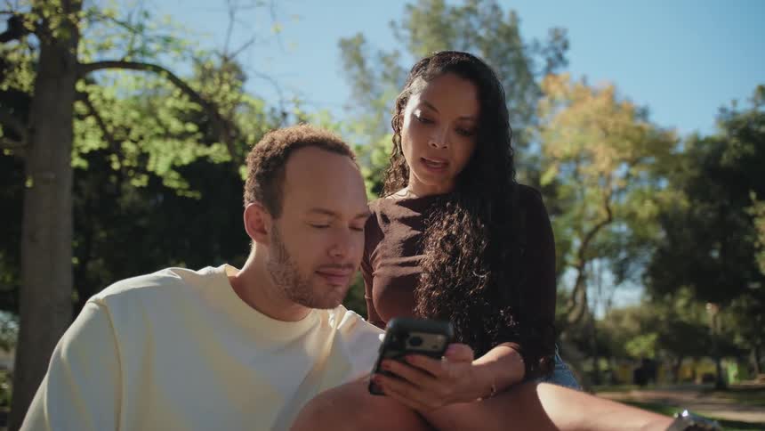 A young couple sits closely together in a sunlit park, looking intently at a smartphone