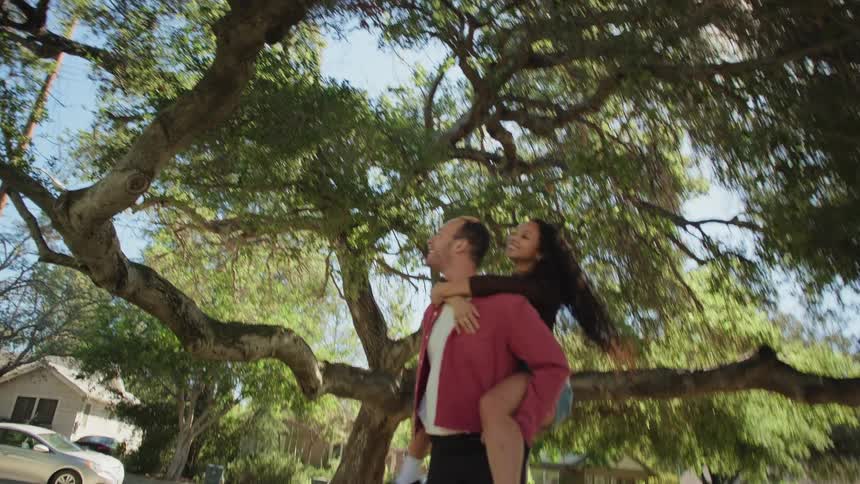 A happy young man gives his girlfriend a piggyback ride as they walk away from the camera under a giant tree on a sunny day