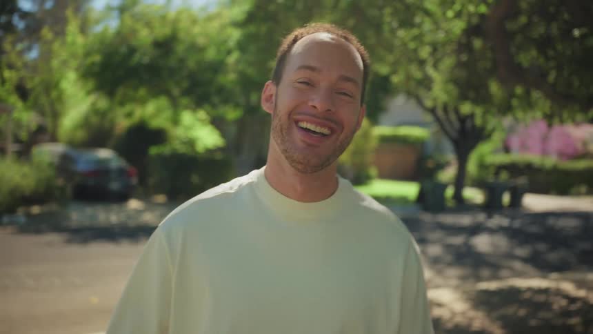 A happy man laughs and playfully leans into the camera for a close-up kiss on a sunny suburban street