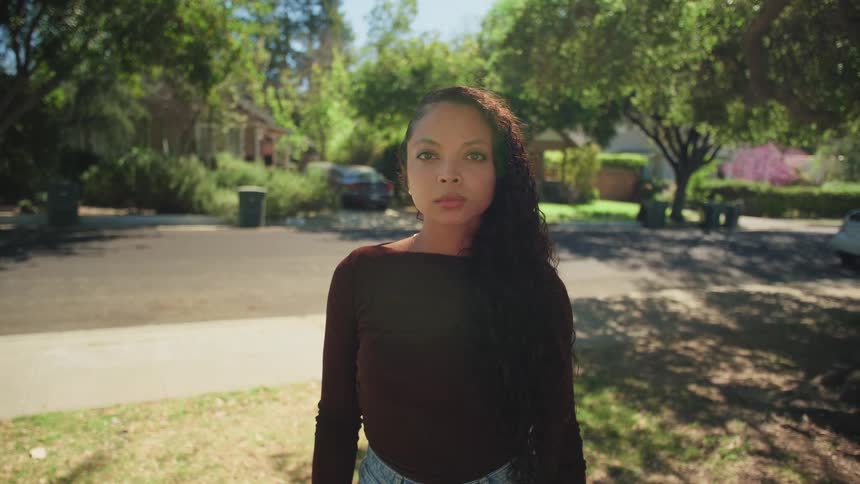 A portrait of a serious young woman with long curly hair standing outdoors in a sunny suburban neighborhood
