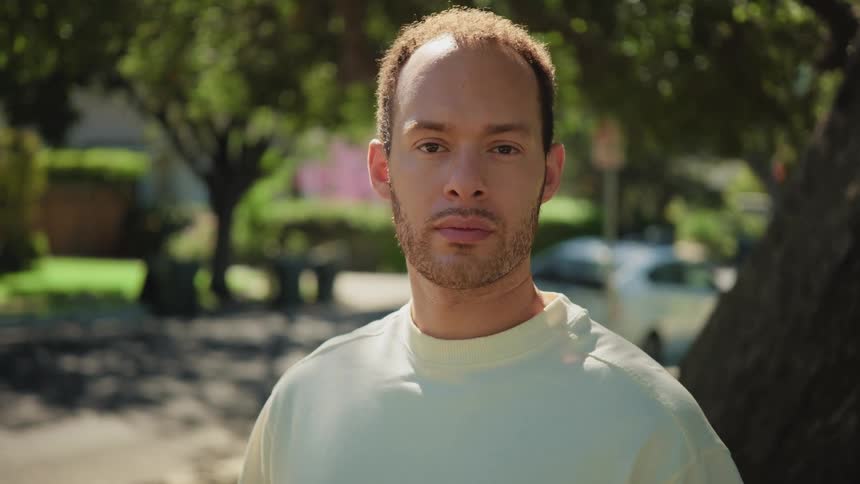Slow zoom-in portrait of a young man with a serious expression standing outdoors on a sunny day