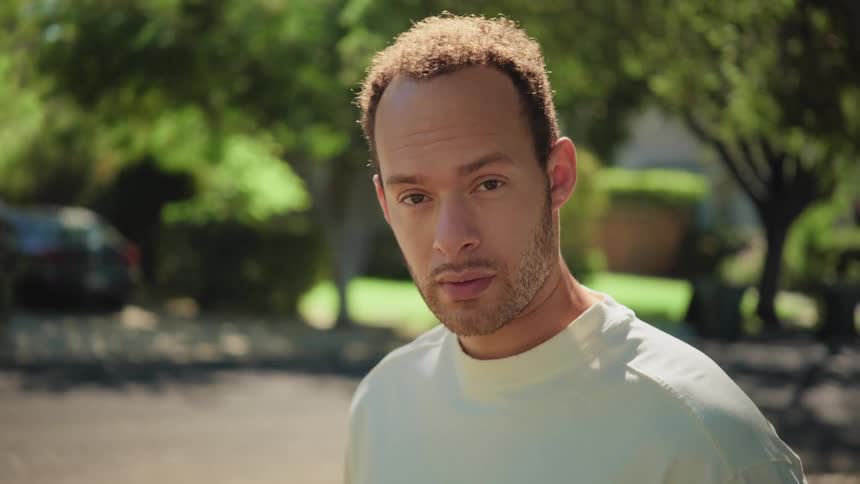 A man with a serious expression looks up and stares into the camera on a sunny suburban street