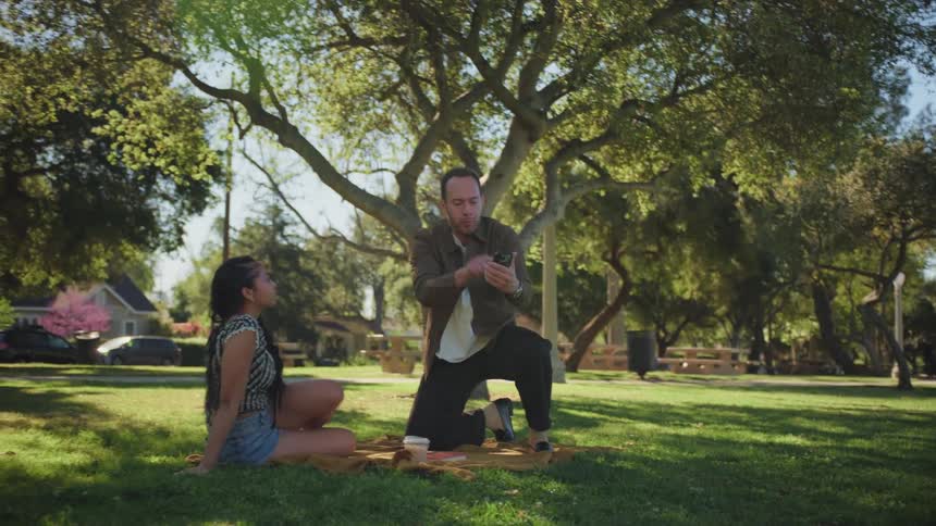 An animated man theatrically performs for a woman as they sit on a picnic blanket in a sunlit park