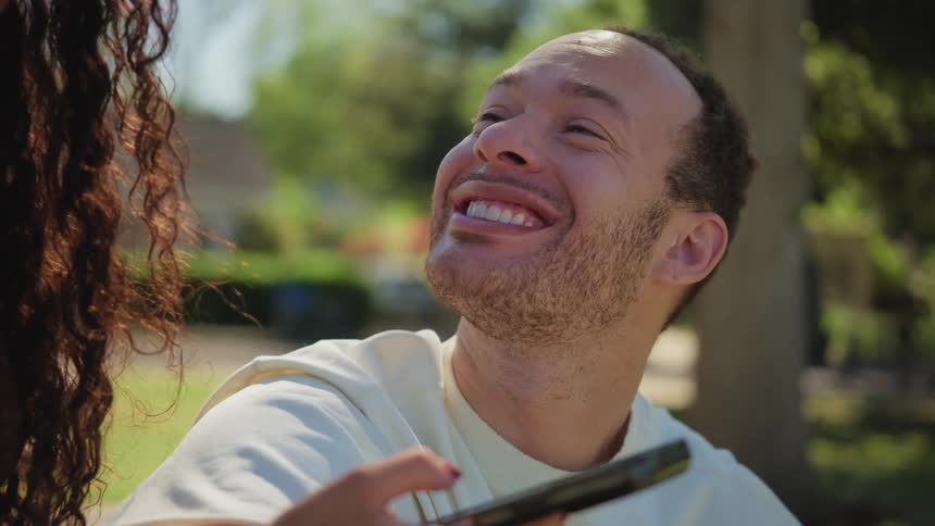 A joyful man bursts into laughter while looking at a phone with a friend in a sunny park in slow motion