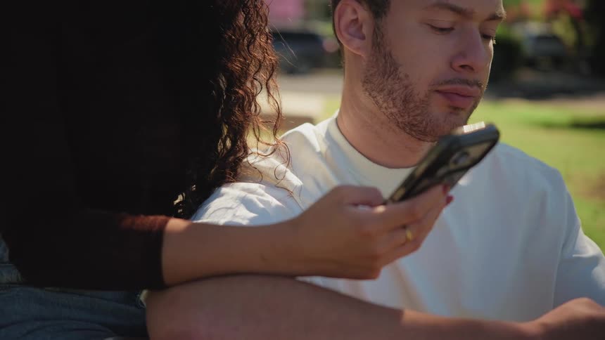 A young woman looks sad and distant while her partner is absorbed in his phone as they sit together in a park