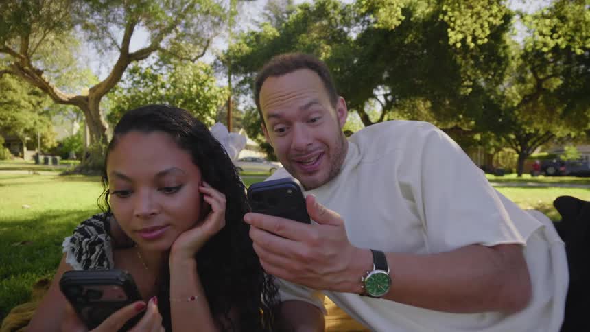 A smiling couple relaxes on a blanket in a park, both engaged with their smartphones on a sunny day as the camera slowly pushes in