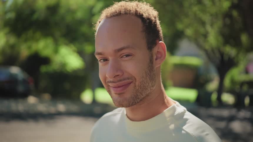 A confident young man looks up and gives a charming smirk to the camera on a sunny day
