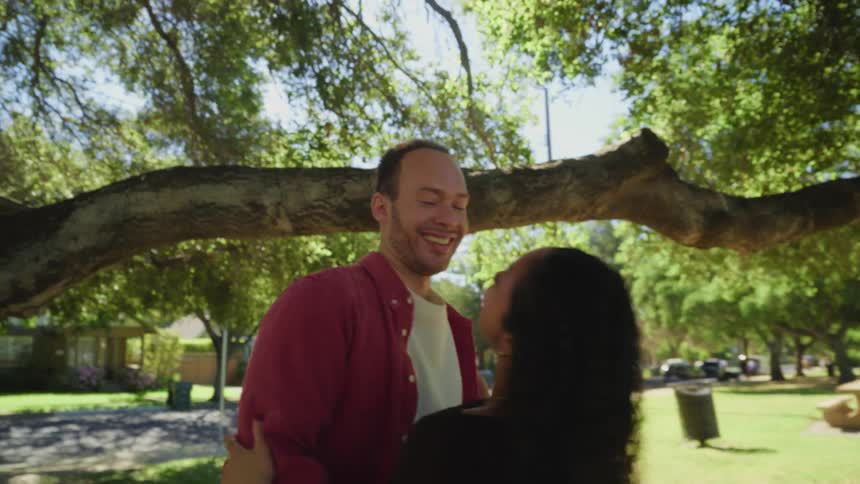 A romantic man dips his laughing partner and pulls her into a loving embrace under a large tree in a sun-drenched park