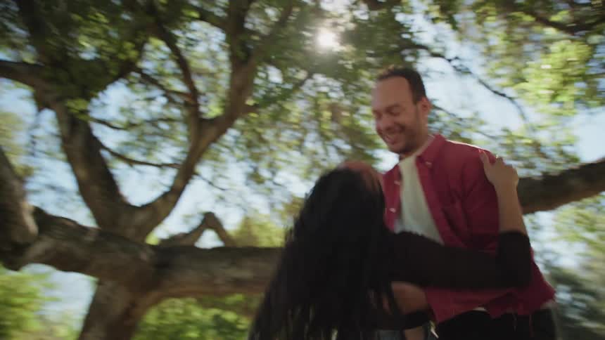A happy man joyfully lifts and spins his girlfriend in his arms as they dance together under a large tree on a sunny day