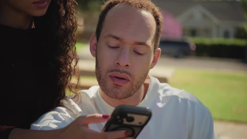 A romantic young couple laughs and shares an intimate kiss while looking at a phone together in a park on a sunny day