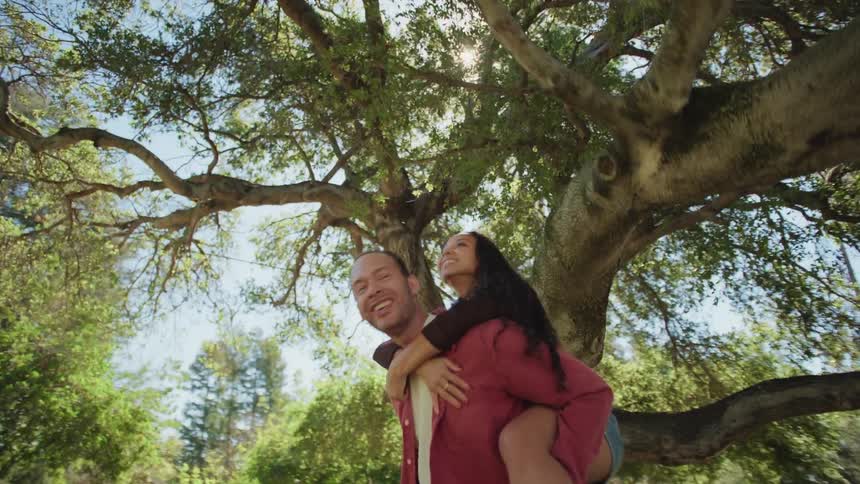 A man gives a woman a piggyback ride under a large tree on a sunny day in slow motion