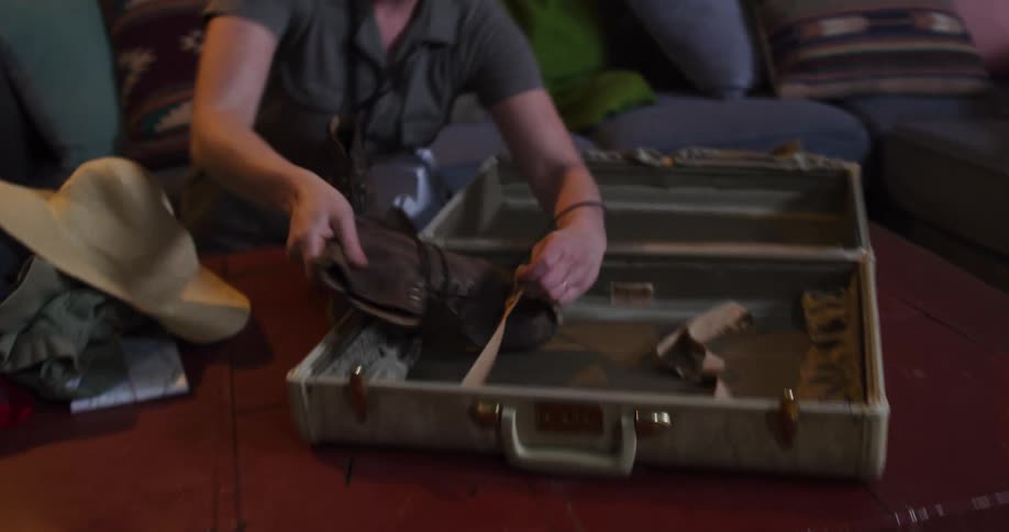 An adventurous woman in slow motion carefully packs her worn leather boots into a vintage suitcase in preparation for a trip