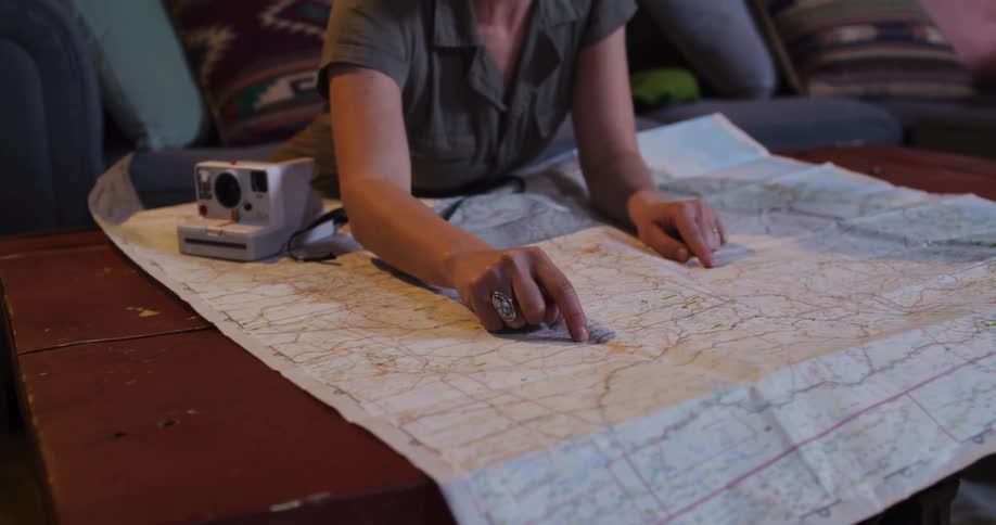 A young woman excitedly plans her next adventure, tracing a route with her finger on a large paper map spread across a coffee table