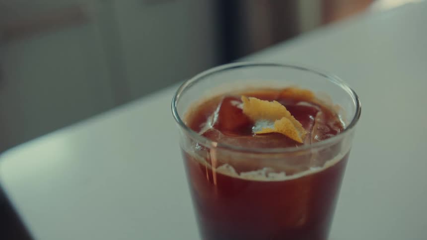 A close-up shot of a glass of refreshing iced coffee with an orange peel garnish sitting on a white table