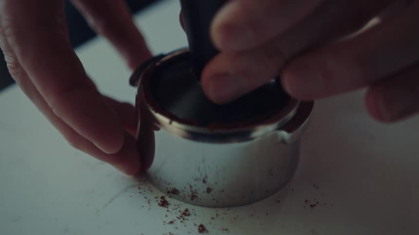 A close-up slow-motion shot of a barista tamping fresh coffee grounds in a portafilter to prepare an espresso