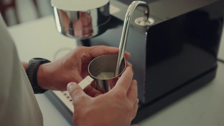 Hands hold a stainless steel jug while an espresso machine's steam wand froths the milk inside