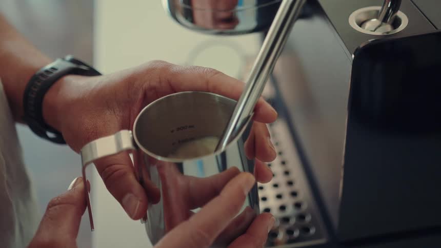 A close-up view of a barista's hands using an espresso machine's steam wand to froth milk in a stainless steel pitcher