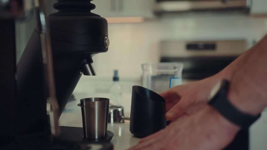 A home barista meticulously prepares espresso by weighing, misting, and grinding fresh coffee beans in a modern kitchen