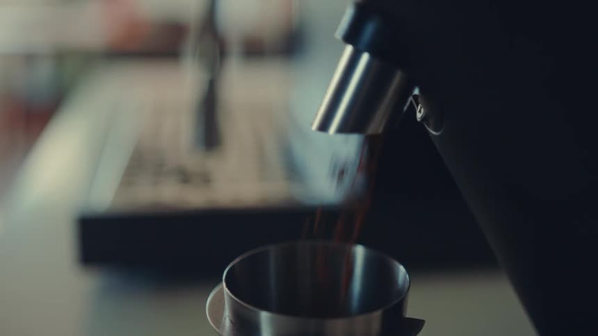 A professional coffee grinder dispenses a stream of freshly ground coffee into a metal cup in cinematic slow motion
