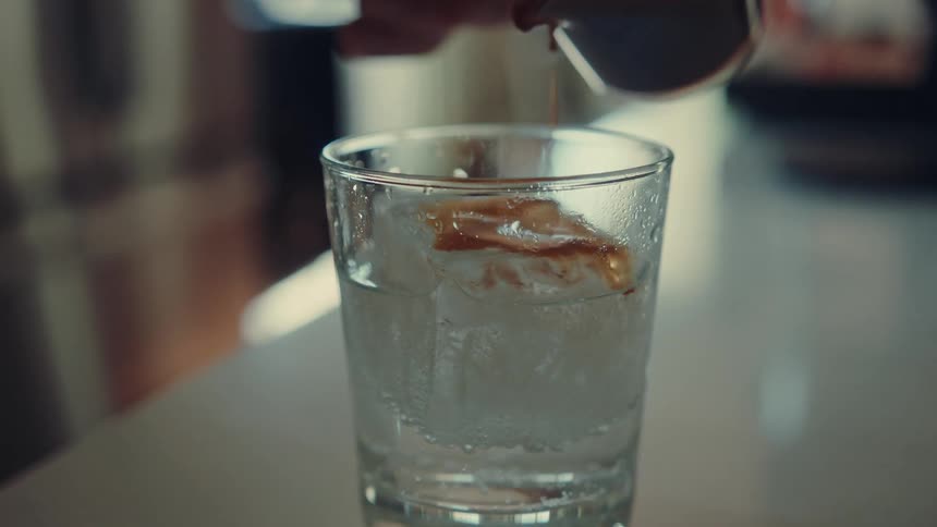 A cinematic slow-motion close-up of an espresso tonic being made as espresso is poured over a large ice cube in a glass of bubbly tonic water