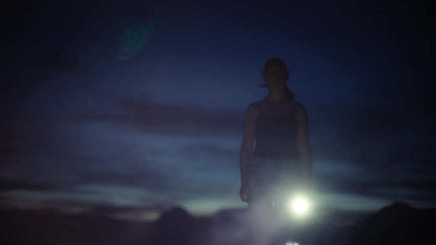 A mysterious woman holding a flashlight stands silhouetted against a dark, cloudy night sky as smoke dissipates around her