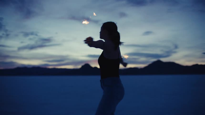 A young woman gracefully spins and dances in slow motion with glowing sparklers at twilight on a vast, open salt flat