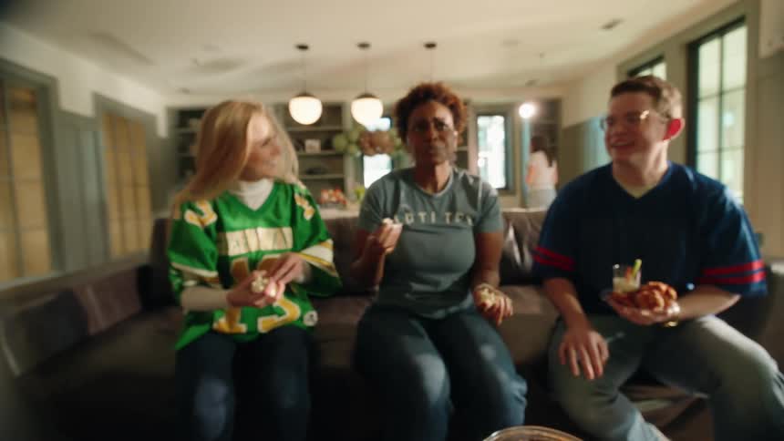 Friends laugh and eat popcorn while watching a football game, with a slow-motion shot of a hand grabbing popcorn from a bowl