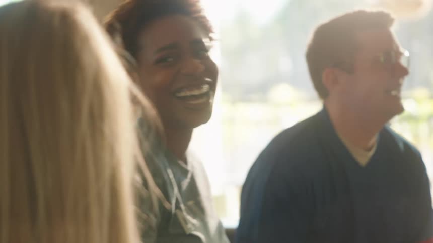A diverse group of friends laugh and cheer in slow motion while watching a game and eating snacks in a sunlit living room