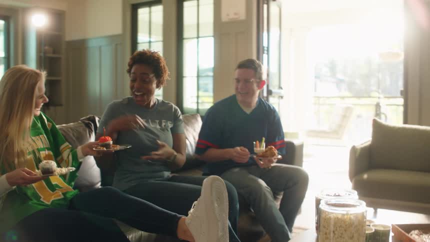 A woman brings cupcakes to her friends as they laugh and talk while watching a game at a party in a sunlit home