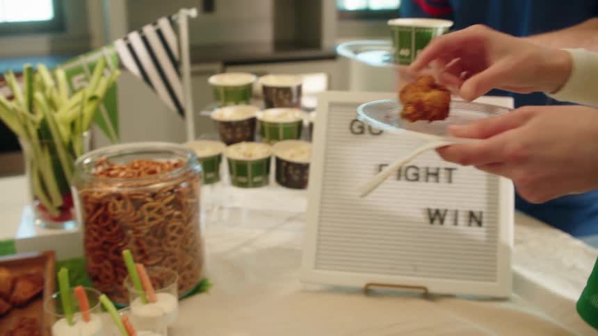 Slow motion close-up of football fans getting wings, pretzels, and dip from a snack table at a game day party