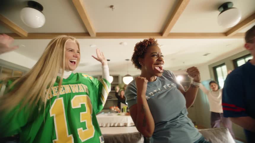 Ecstatic friends leap up from the couch and celebrate wildly in slow motion while watching a sports game during a lively house party
