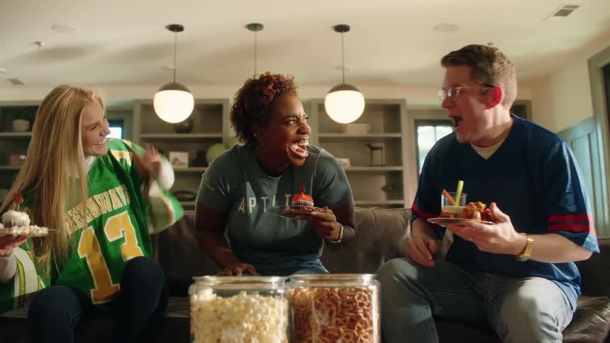 Three diverse friends in football jerseys cheer and laugh while watching a sports game together on the couch