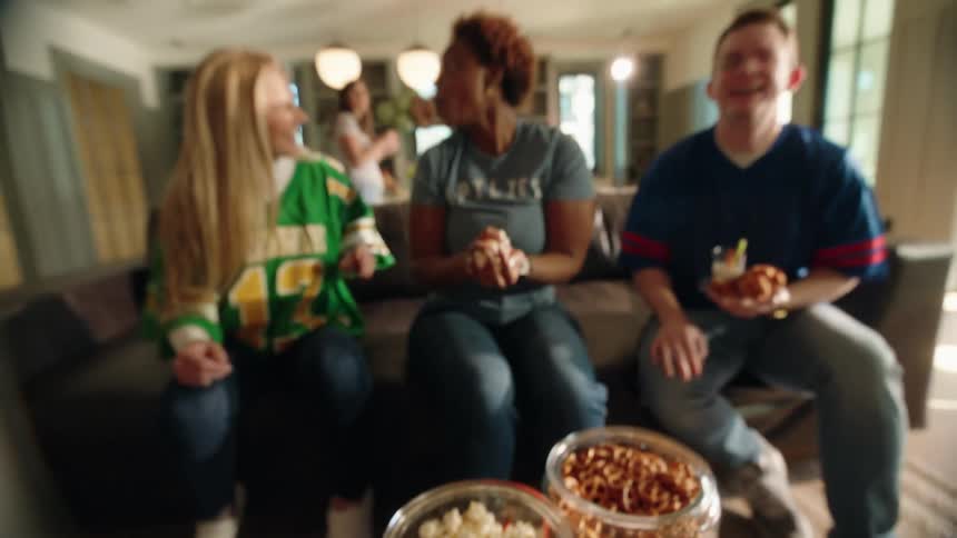 A close-up of a hand grabbing popcorn from a bowl pulls back to reveal a group of friends laughing while watching a sports game at a party