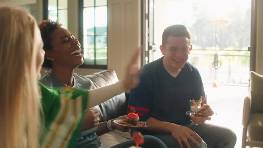 Excited diverse friends laugh and high-five each other while celebrating a game-day victory with cupcakes and wings