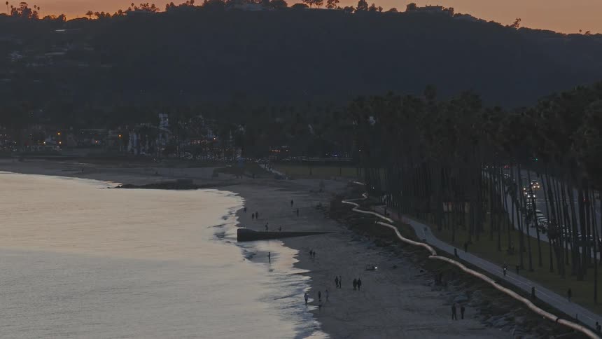 A scenic wide shot of a California beach at dusk as people walk along the shore and cars drive on the adjacent road lined with palm trees