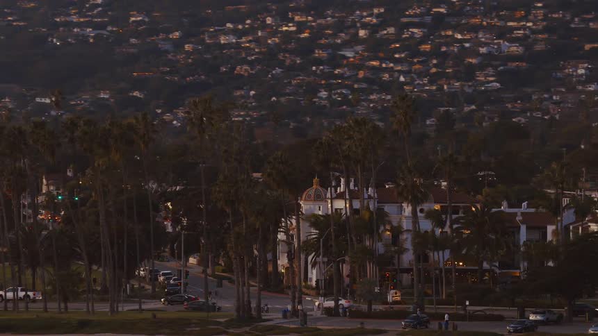A slow pan reveals a picturesque view of Santa Barbara's coastline street, lined with palm trees and Spanish-style architecture, as twilight settles over the mountains