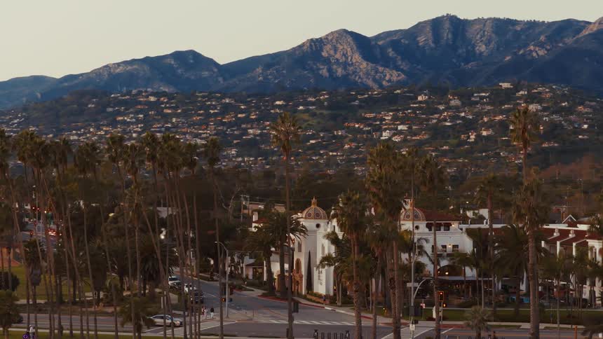 A slow zoom out reveals the scenic coastal city of Santa Barbara, nestled between rows of palm trees and the Santa Ynez mountains at sunset