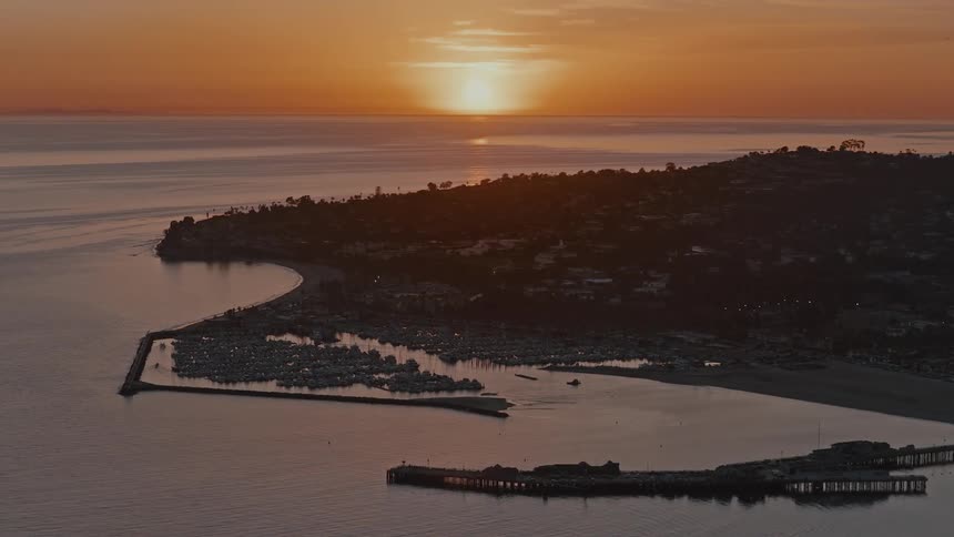 A serene wide shot captures the stunning orange sunset over the calm ocean, with a pier and a boat-filled marina silhouetted along the coastline