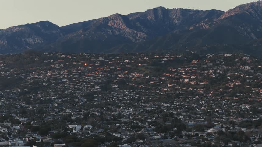 A timelapse of lights illuminating homes in a sprawling hillside community at the base of a mountain range as evening arrives