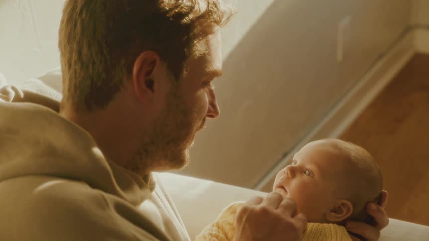 A father lovingly gazes at his newborn baby, gently stroking their face, causing the infant to smile in the warm, golden sunlight