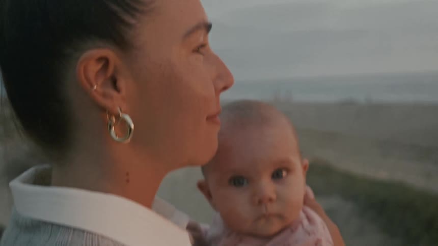 A mother tenderly holds her baby daughter while overlooking a sandy beach at twilight, kissing her on the forehead