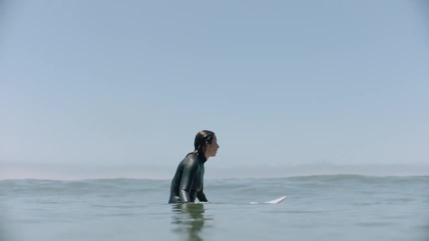 A serene, water-level view of a female surfer sitting on her board, waiting for waves before the camera dips underwater