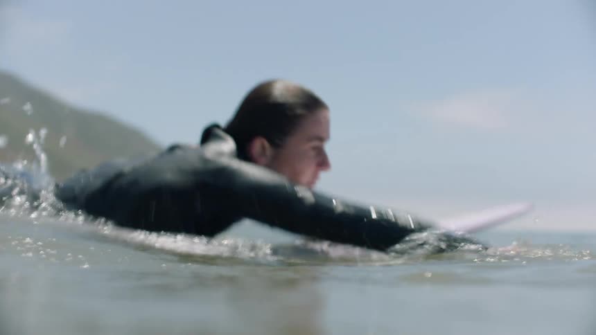 An athletic woman in a wetsuit paddles energetically on her surfboard in the ocean in a dynamic handheld shot