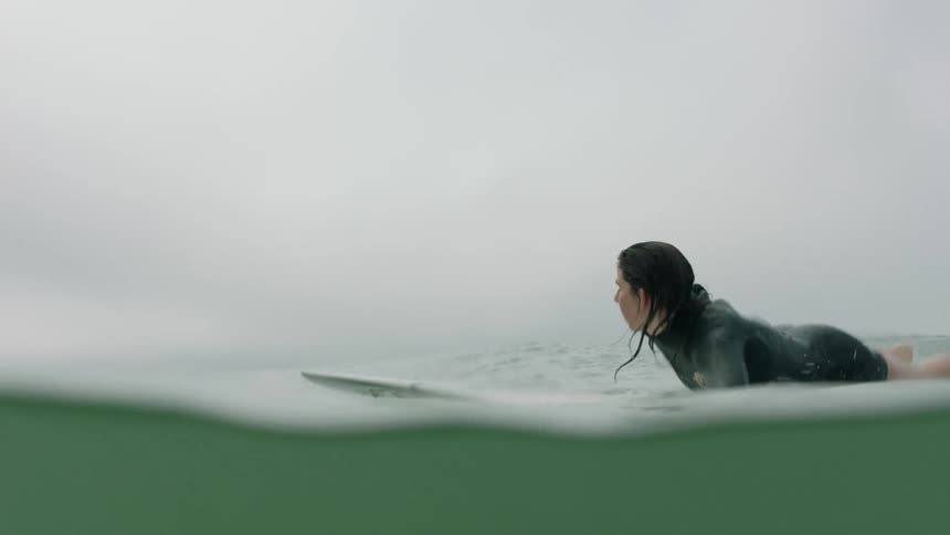 A woman in a wetsuit paddles her surfboard across the calm ocean on a summer day, shot in slow motion from a water-level perspective